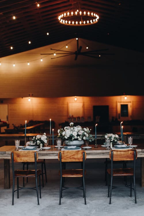 wooden table and chairs set up for wedding reception inside tall modern barn with chandeliers